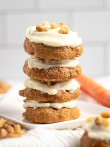 Four beautifully stacked carrot cake cookies frosted with cream cheese frosting and sprinkled with chopped walnuts surrounded by chopped walnuts and an uncut carrot in the background.