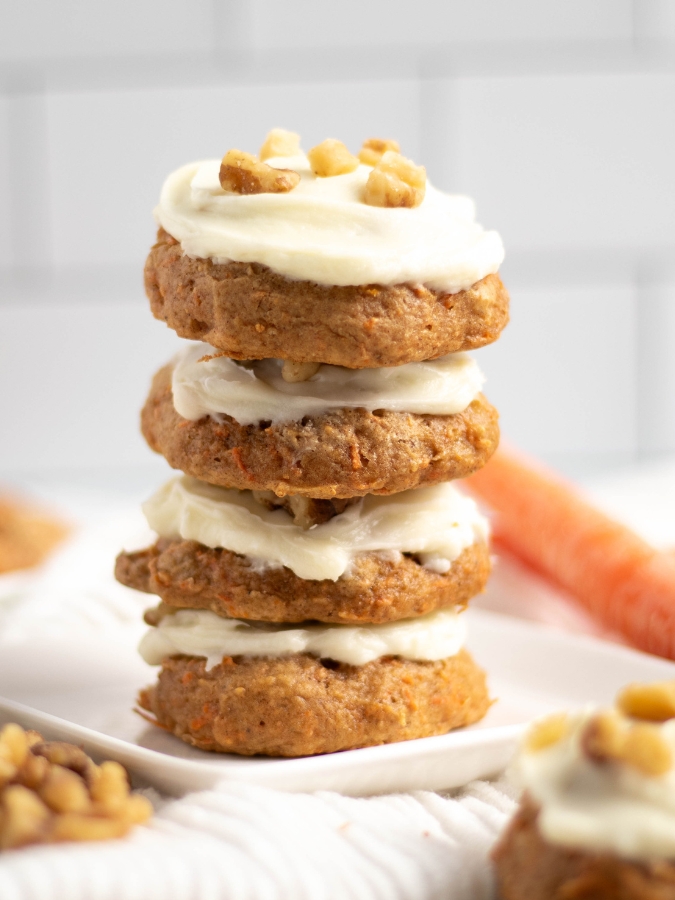 Four beautifully stacked carrot cake cookies frosted with cream cheese frosting and sprinkled with chopped walnuts surrounded by chopped walnuts and an uncut carrot in the background.