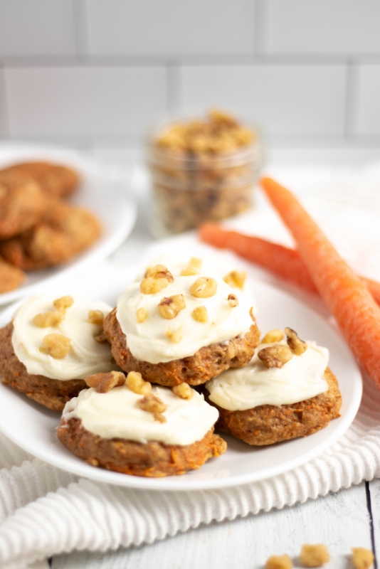 A plate of 5 carrot cake cookies frosted with cream cheese frosting and carrots, walnuts and unfrosted cookies in the background.