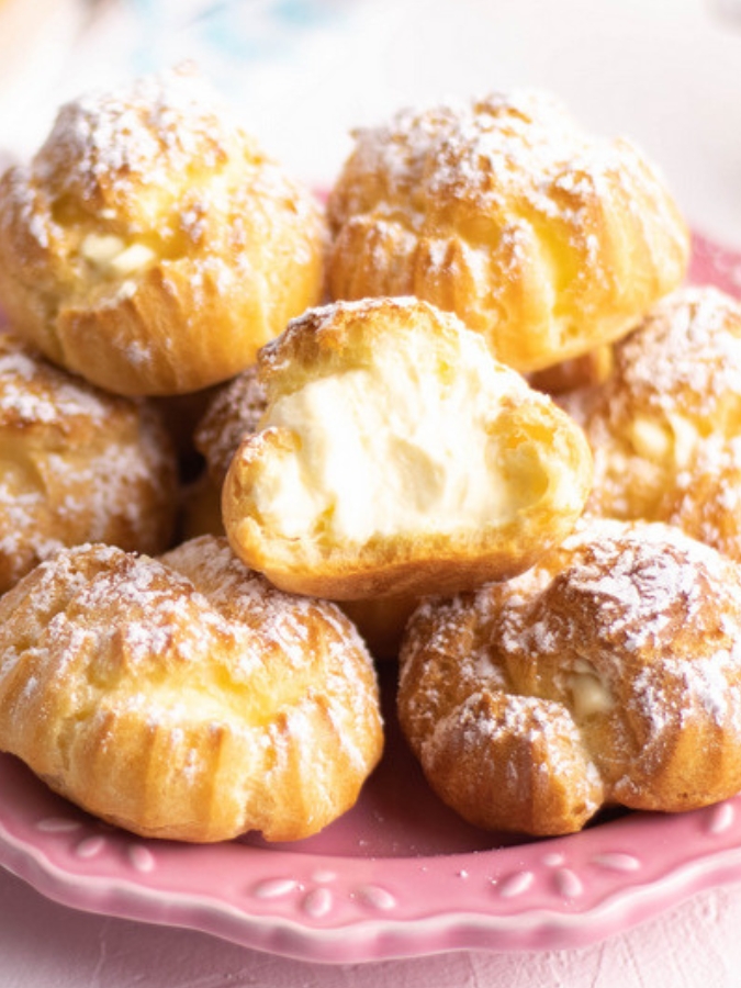 A pile of perfectly baked cream puffs with the center cream puff cut and showing the filling.