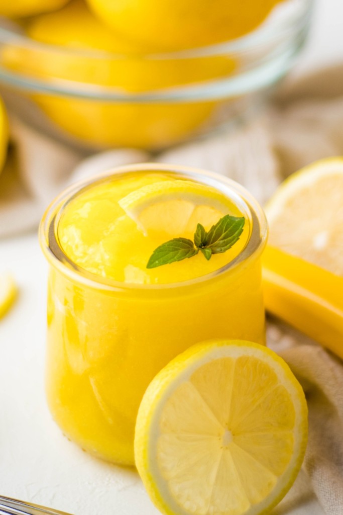 Small glass cup of bright yellow lemon curd garnished with a mint leaf and slice of lemon.