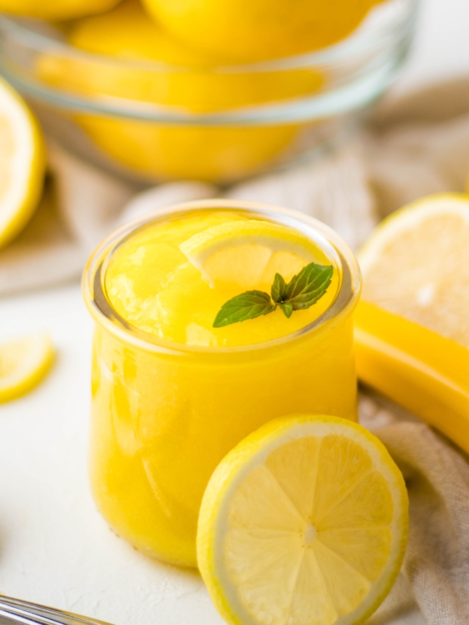 Small glass cup of bright yellow homemade lemon curd garnished with a mint leaf and slice of lemon.