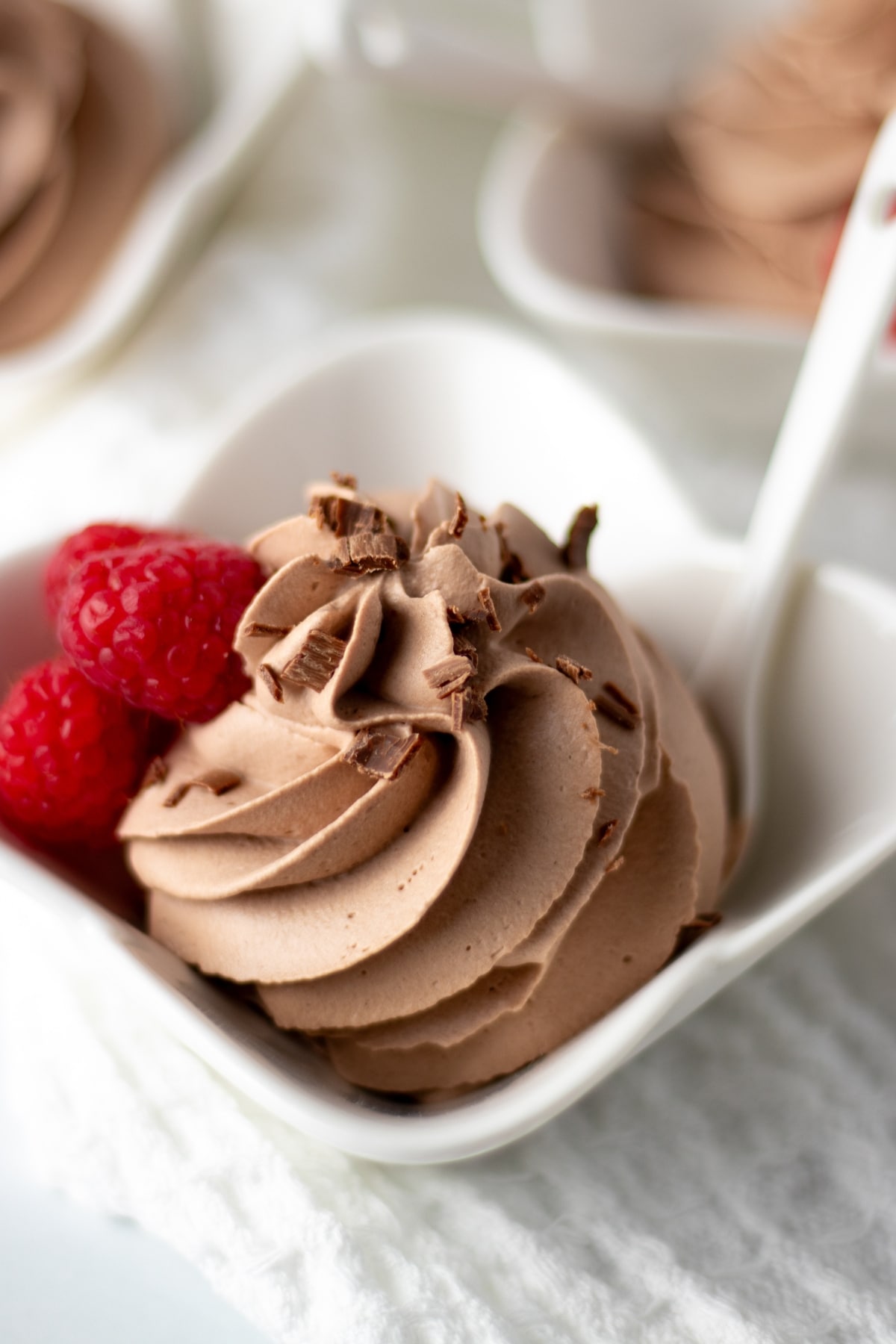 A small dish with a swirl of chocolate mousse sprinkled with chocolate and garnished with 3 red raspberries.