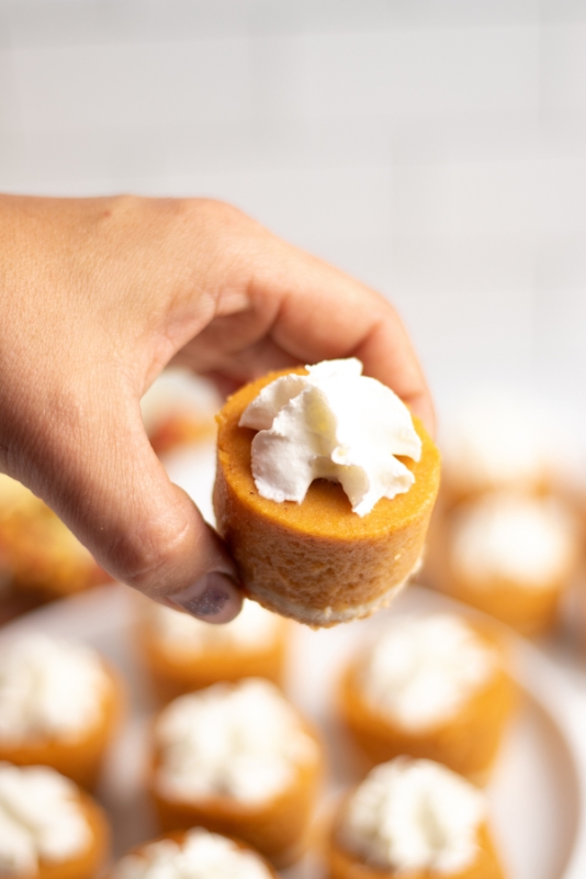A hand holding a mini pumpkin pie bite with more pies in the background.